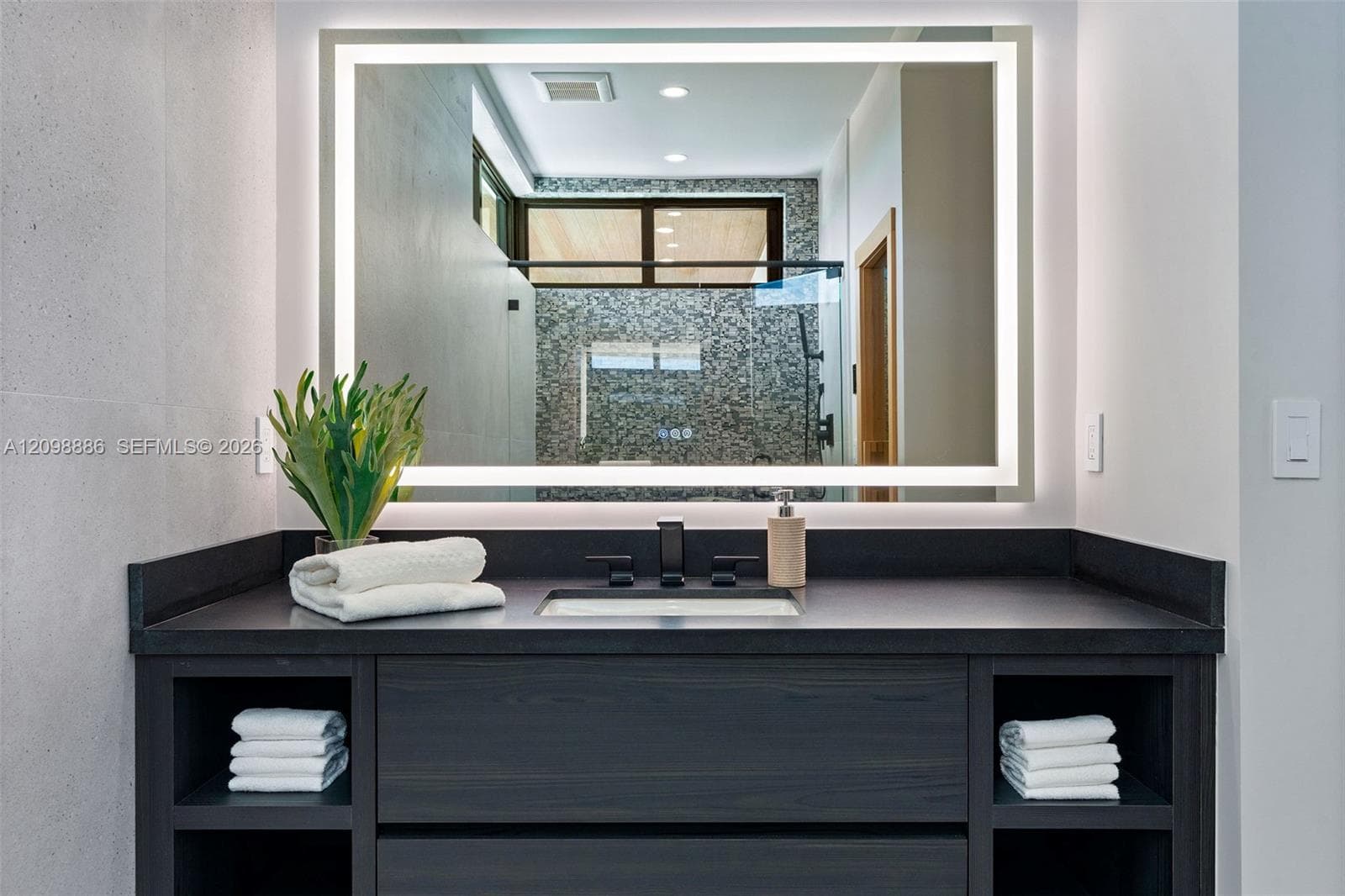 Dark wood vanity featuring integrated open shelving, a dark solid-surface countertop, an undermount rectangular sink, and a matte black faucet paired with a rectangular LED-backlit mirror
