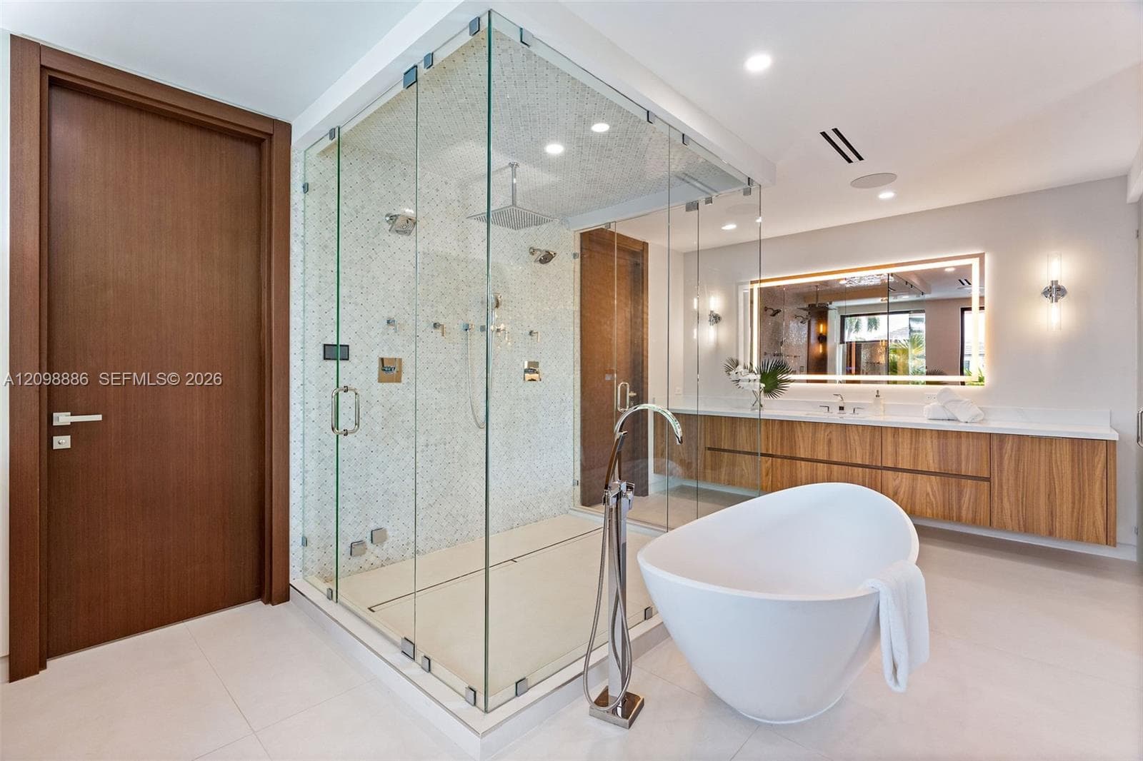 Glass-enclosed walk-in shower with mosaic tile surround and rainfall showerhead, freestanding soaking tub with floor-mounted faucet, wall-mounted wood-finish vanity with countertop, and integrated LED-backlit mirror