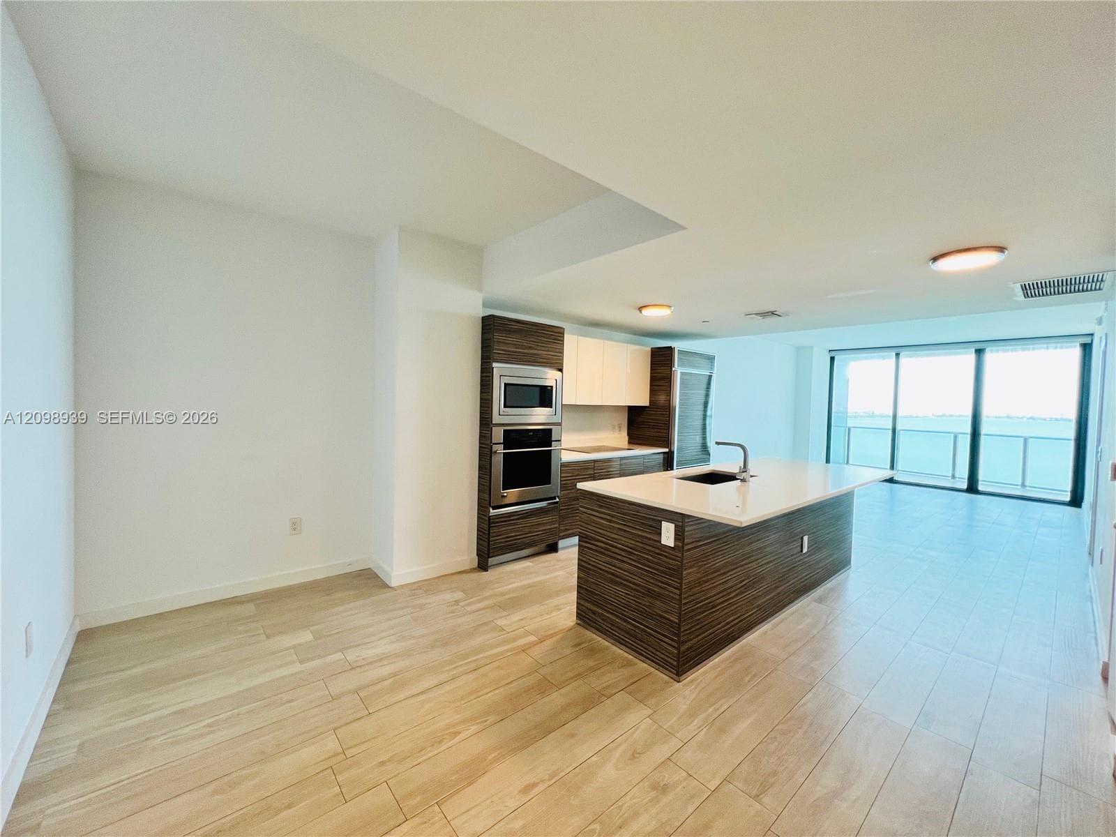 Wood-finish flooring throughout, full-height sliding glass doors, white walls, and a kitchen featuring an island with an undermount sink and stainless steel appliances