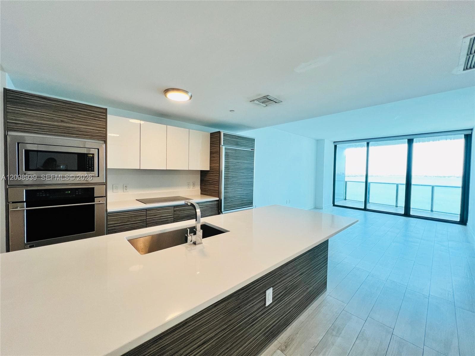 Open-concept kitchen featuring an oversized island with an integrated stainless steel sink, stainless steel appliances, wood-finish cabinetry, and white upper cabinets