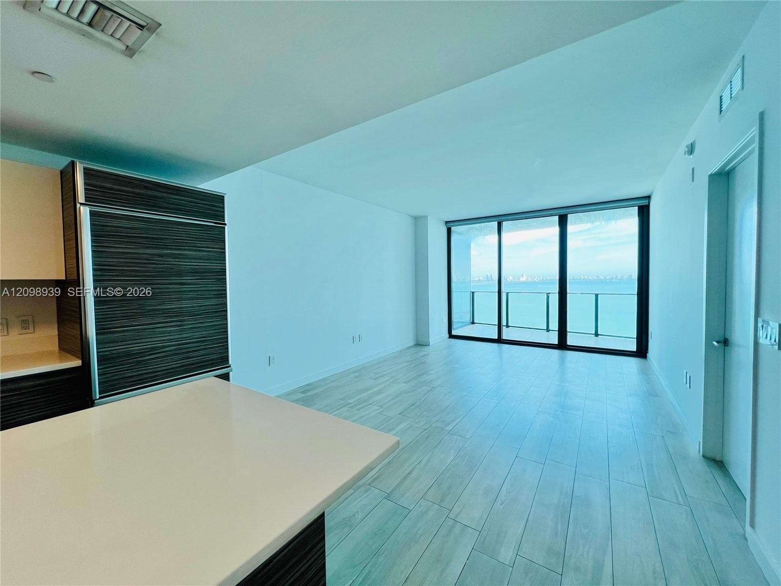 Open-concept living area with floor-to-ceiling sliding glass doors, a built-in paneled refrigerator, light-toned wood-finish flooring, and a stone countertop island