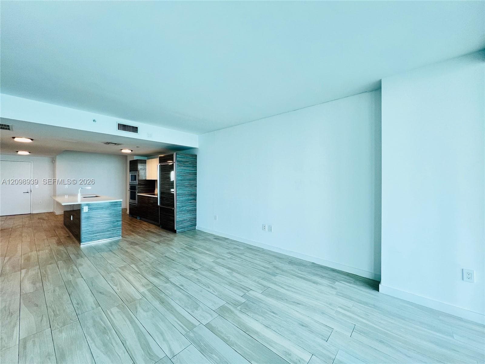 Wood-finish flooring throughout, modern kitchen island with white countertop, integrated stainless steel appliances, and ceiling-mounted recessed lighting