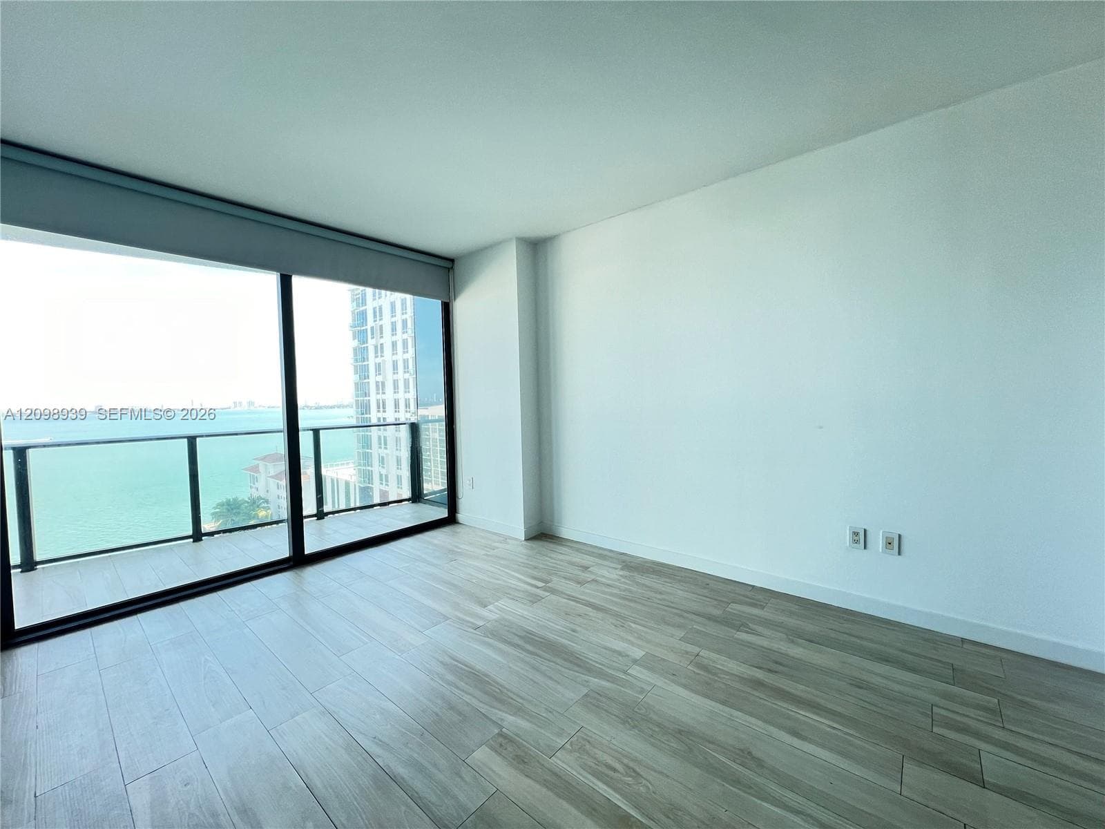 Floor-to-ceiling sliding glass doors, balcony with glass railing, wood-finish flooring, white walls, and roller shades