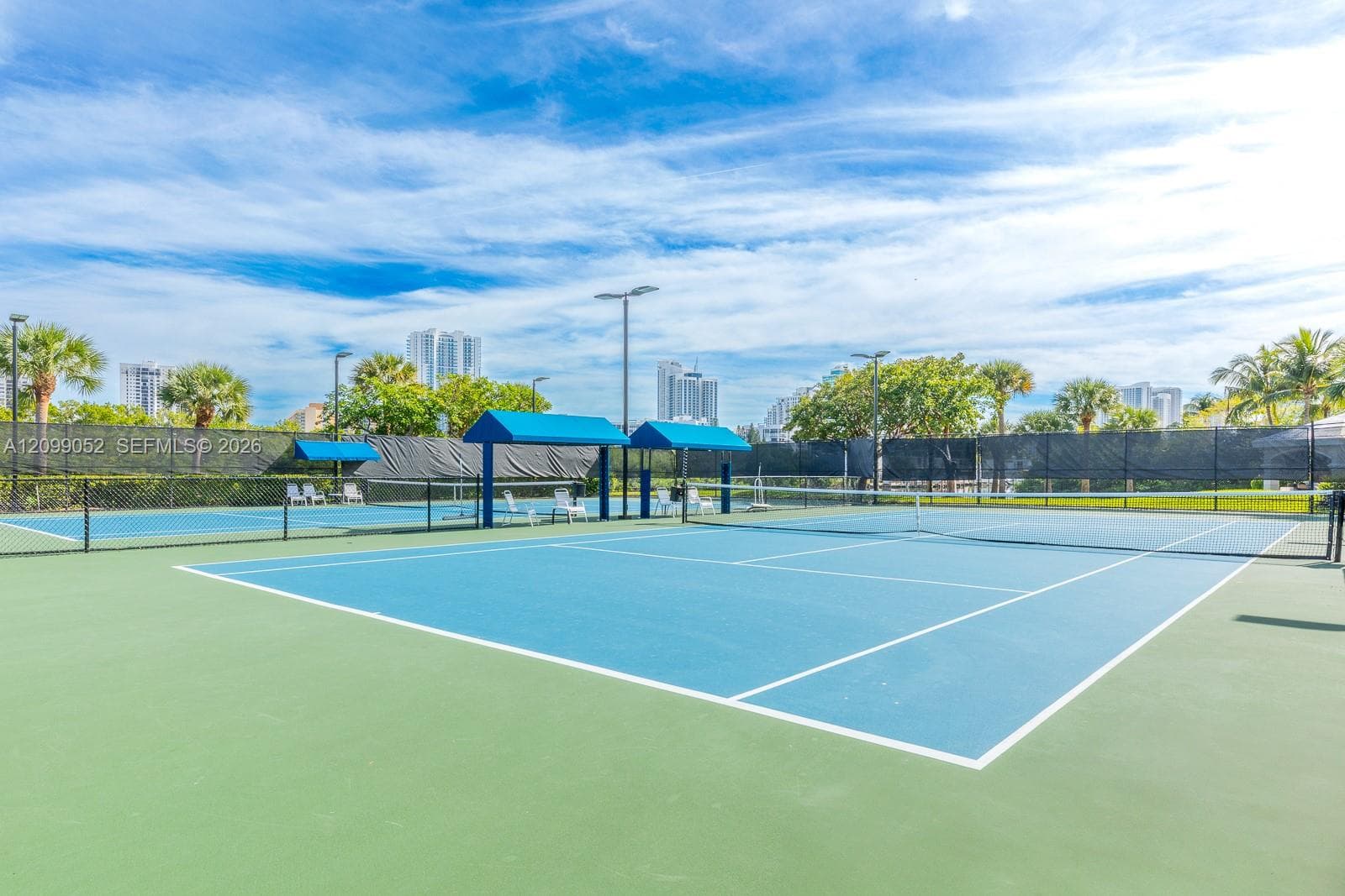 Hard-surface tennis courts featuring perimeter mesh fencing, blue sunshades, integrated court lighting, and mature landscaping