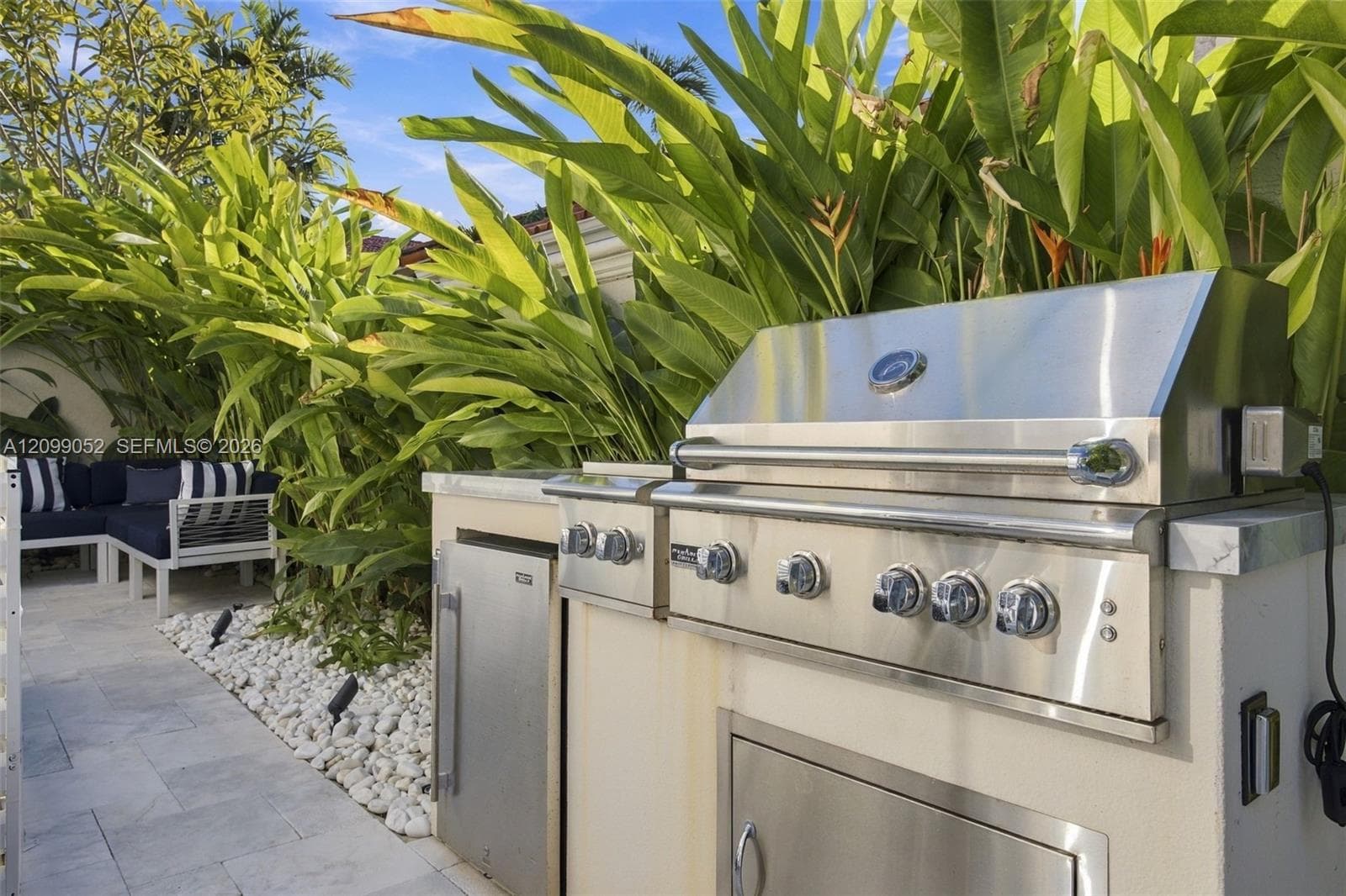 Outdoor kitchen featuring a stainless steel grill, stone-surfaced countertops, custom built-in cabinetry, and integrated storage, set against a landscaped garden bed