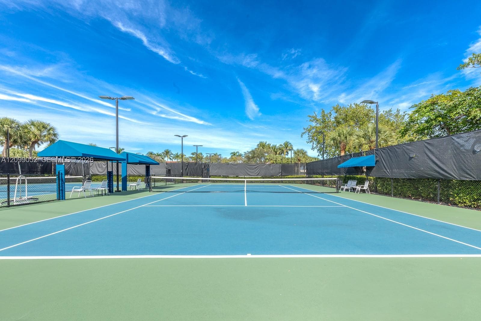 Hard-court tennis facility featuring blue and green surface markings, perimeter privacy fencing, overhead lighting, and built-in shaded seating areas