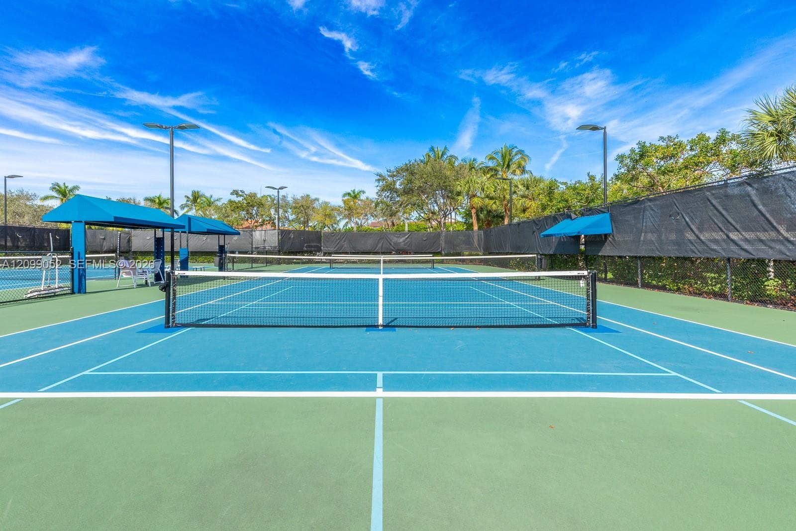 Hard-court tennis facility featuring blue and green surface, permanent net, protective perimeter fencing, pole-mounted lighting, and integrated shaded structures