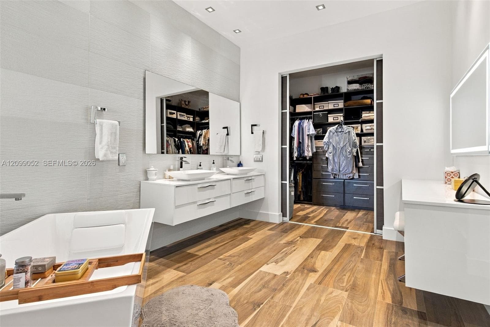 Floating white dual-sink vanity with rectangular vessel sinks, freestanding deep-soaking tub, wood-finish flooring, and built-in closet system with sliding glass-panel doors