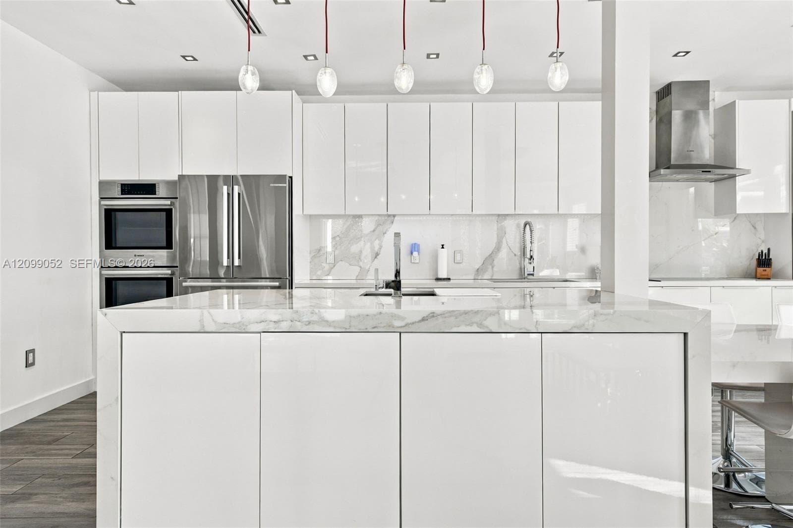 Kitchen featuring high-gloss white cabinetry, a waterfall-edge stone island, stainless steel built-in double wall ovens, a coordinating range hood, and pendant lighting