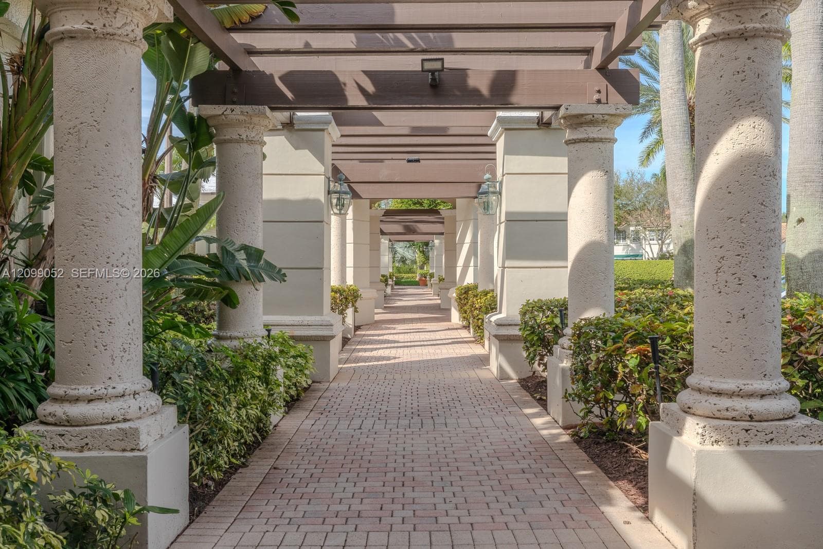 Outdoor walkway featuring a brick-paver path, structural stone columns, overhead timber-frame pergola, and integrated wall-mounted lantern lighting