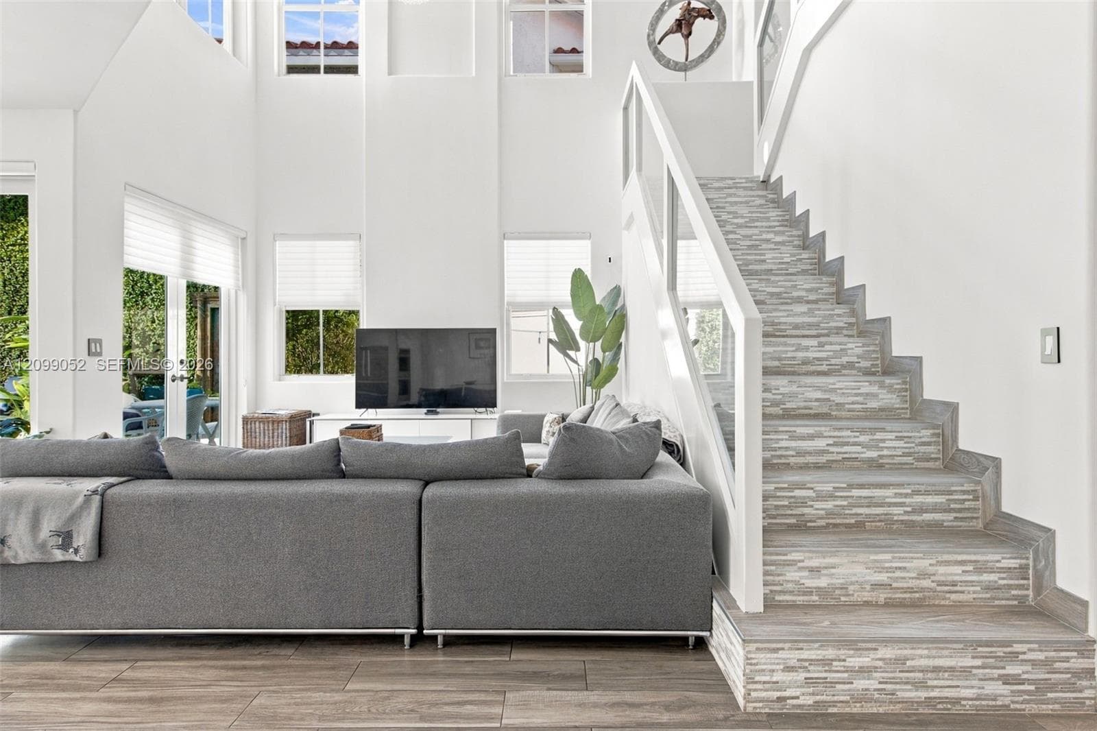 Double-height ceiling, open floor plan featuring wood-finish flooring, staircase with tiled risers, white railing, and glass paneled walls, and multiple large windows with cellular shades
