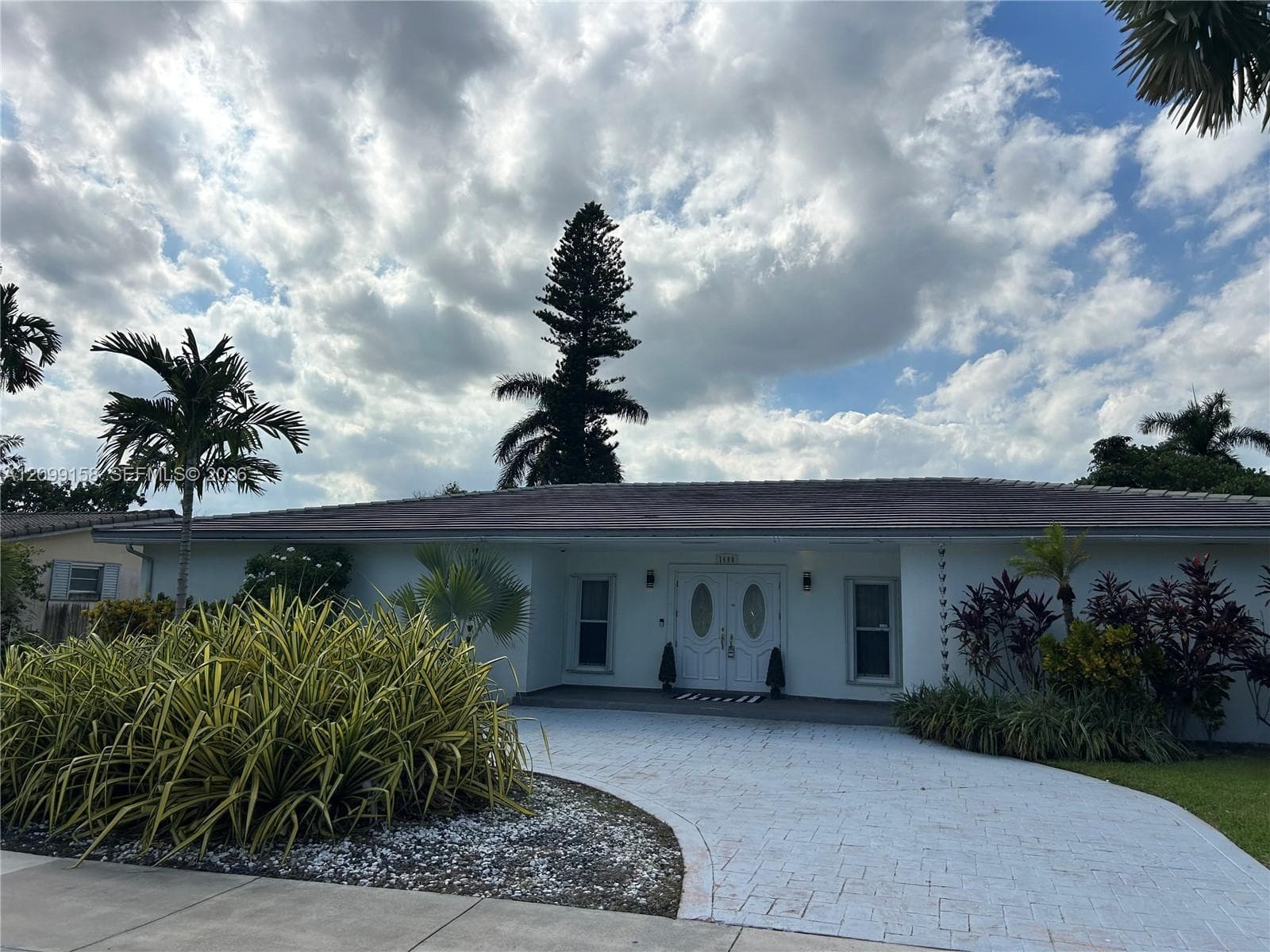Single-story exterior featuring a tile roof, paved circular driveway, double-door entry with glass inserts, and established tropical landscaping