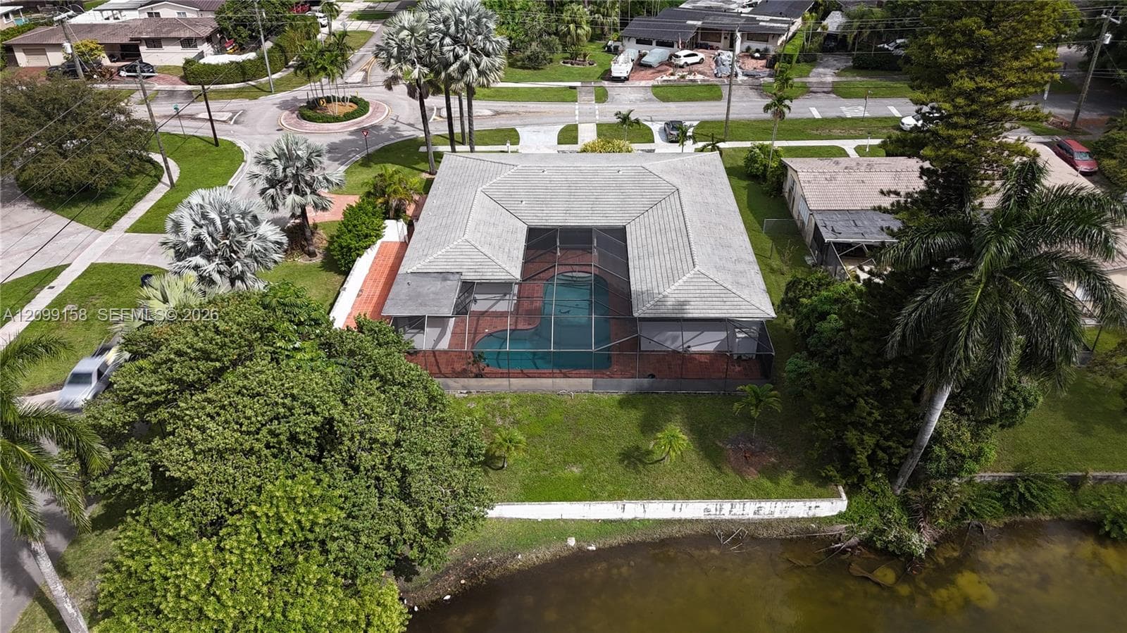 Aerial perspective of a single-story residence featuring a screened-in pool enclosure, tiled patio, concrete tile roof, and waterfront location with mature landscaping