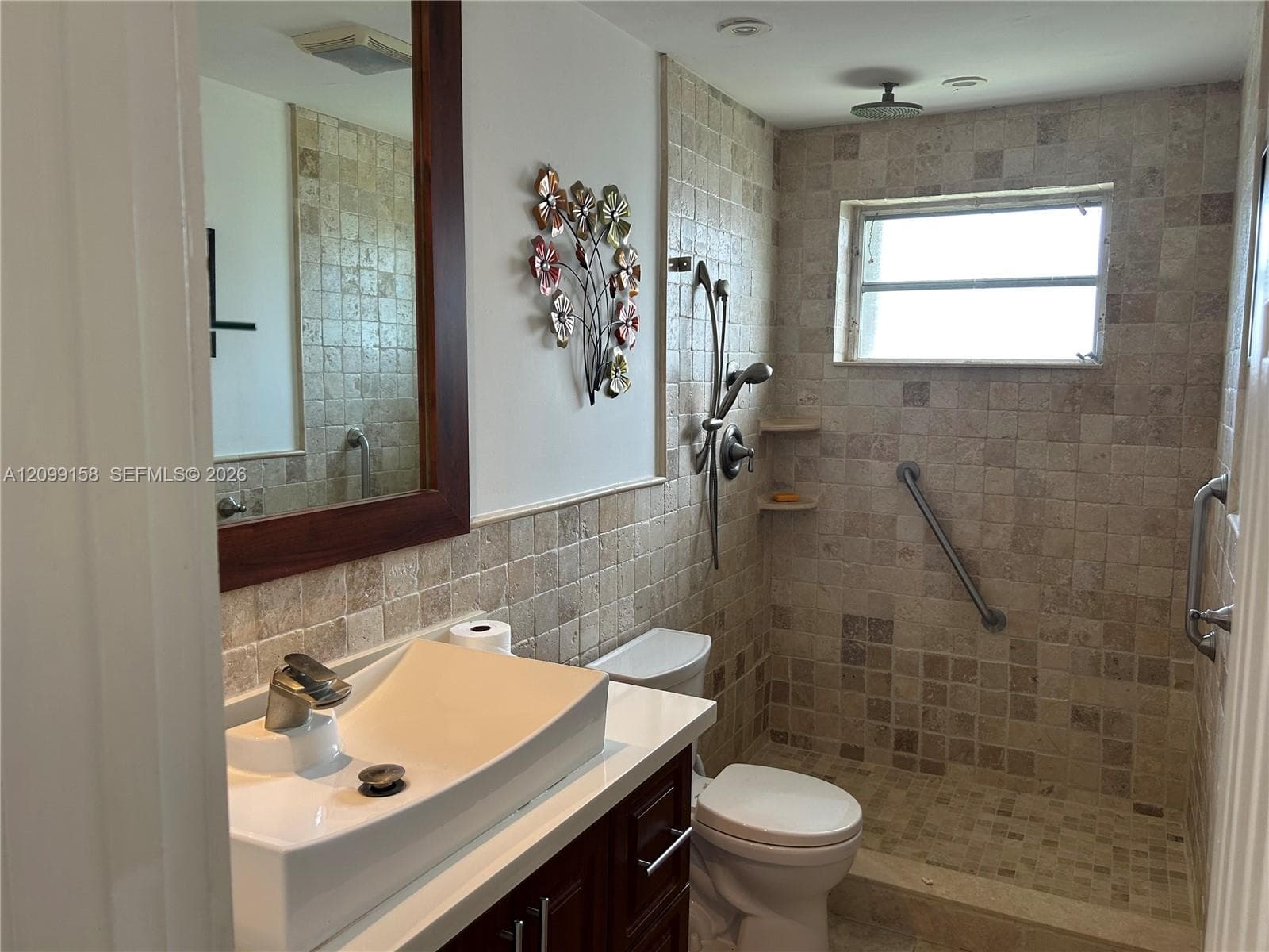 Bathroom featuring square stone tile wall and floor surfaces, vessel sink with chrome faucet, dark wood-finish vanity, and walk-in shower with rain showerhead