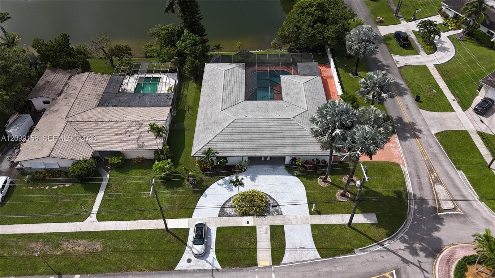 Aerial perspective of waterfront property featuring a U-shaped home, screened-in pool enclosure, circular concrete driveway, and mature landscaping
