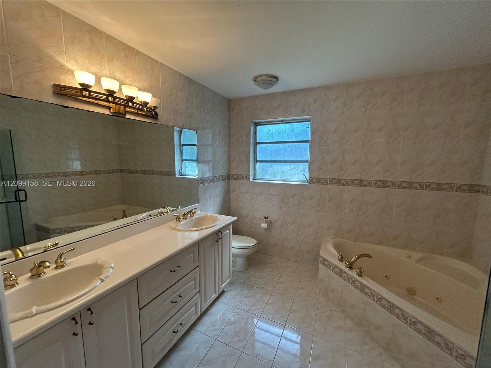 Dual-sink vanity with solid-surface countertop and metallic-finish faucets, large wall-mounted mirror, ceramic tile wall and floor surfaces, and built-in jetted soaking tub