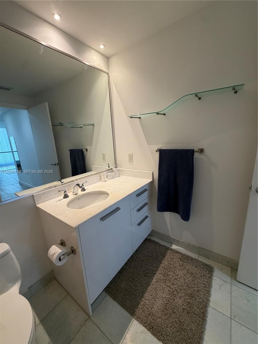 Bathroom featuring a white vanity cabinet, light-toned stone-finish countertop, undermount sink with chrome-finish widespread faucet, wall-spanning mirror, and floating glass shelving