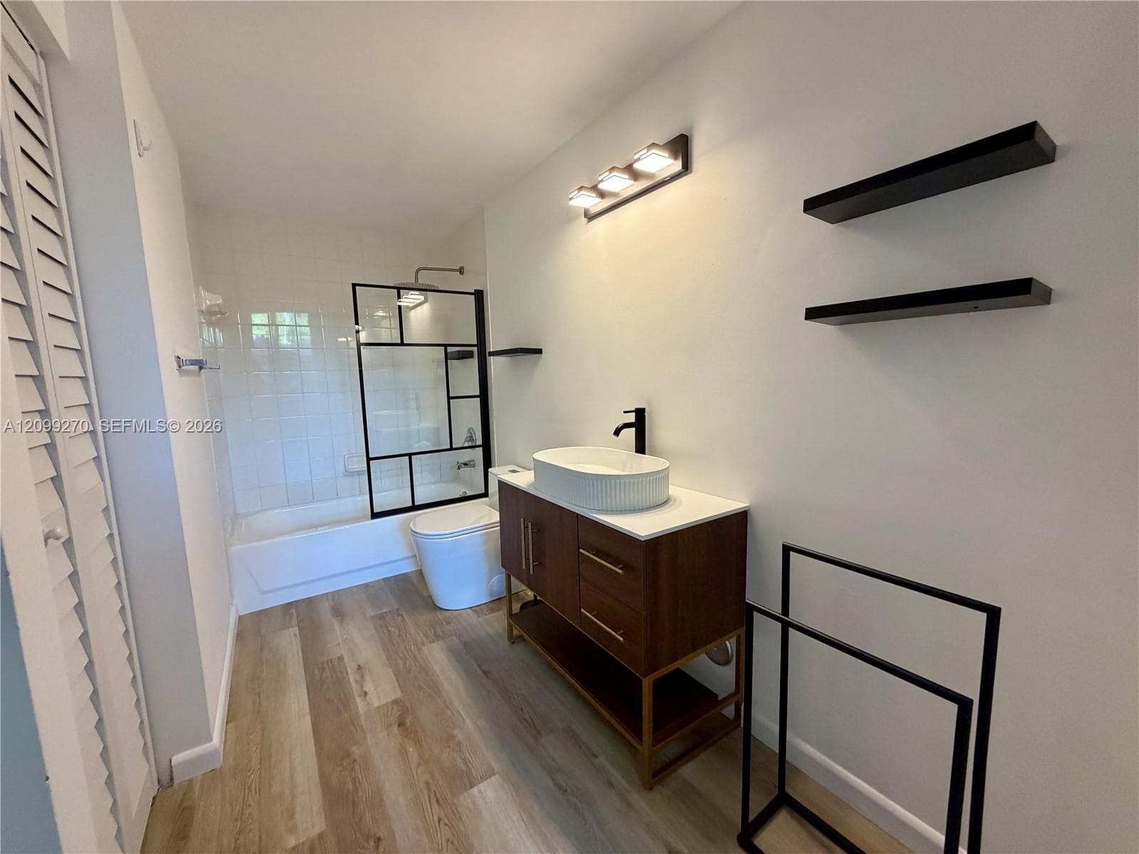 Bathroom featuring a vanity with a white vessel sink, wood-finish cabinetry, matte black hardware, a shower enclosure with a geometric frame, and wood-finish flooring