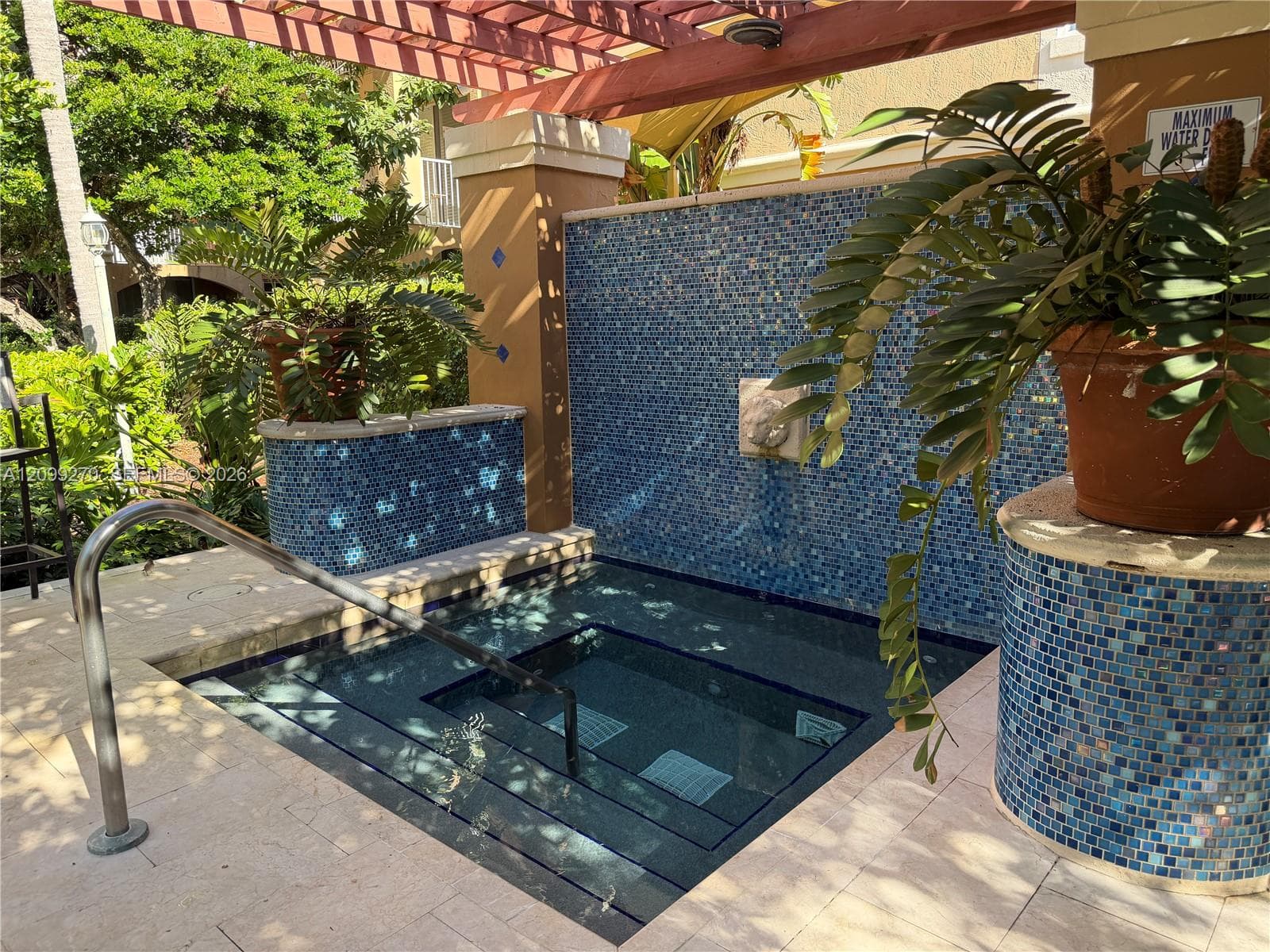 In-ground spa featuring a blue mosaic tile water feature, stone-finished decking, and a wood-slat overhead trellis