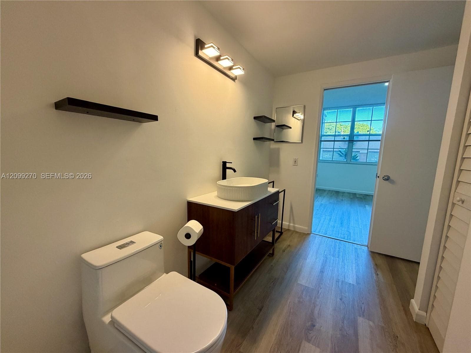 Bathroom featuring a vanity with a white vessel sink, matte black faucet, wall-mounted shelving, wood-finish flooring, and a modern three-light fixture