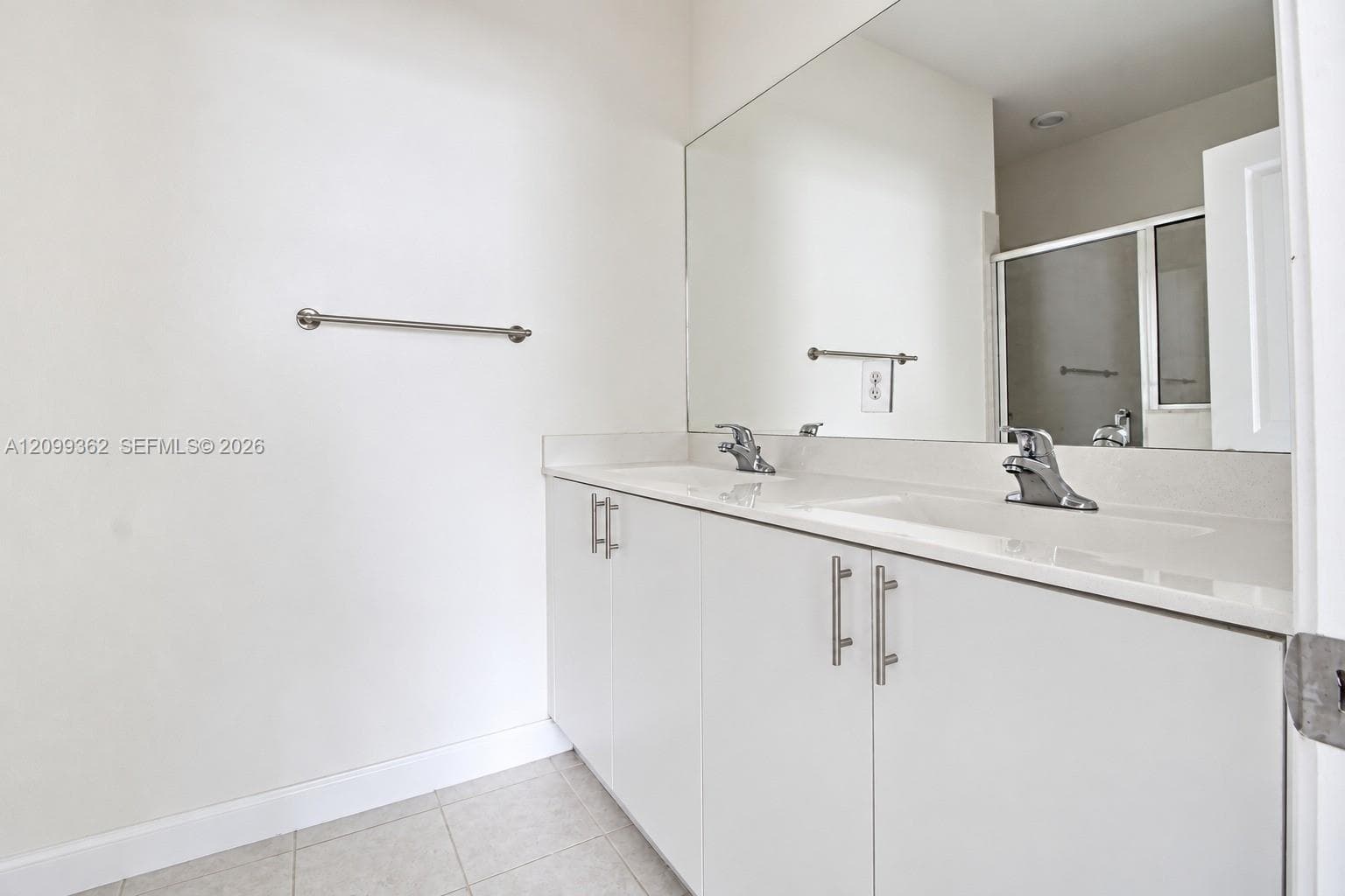 Dual-sink vanity with a solid-surface countertop, cabinetry featuring stainless steel pulls, an expansive wall-mounted mirror, and tile flooring