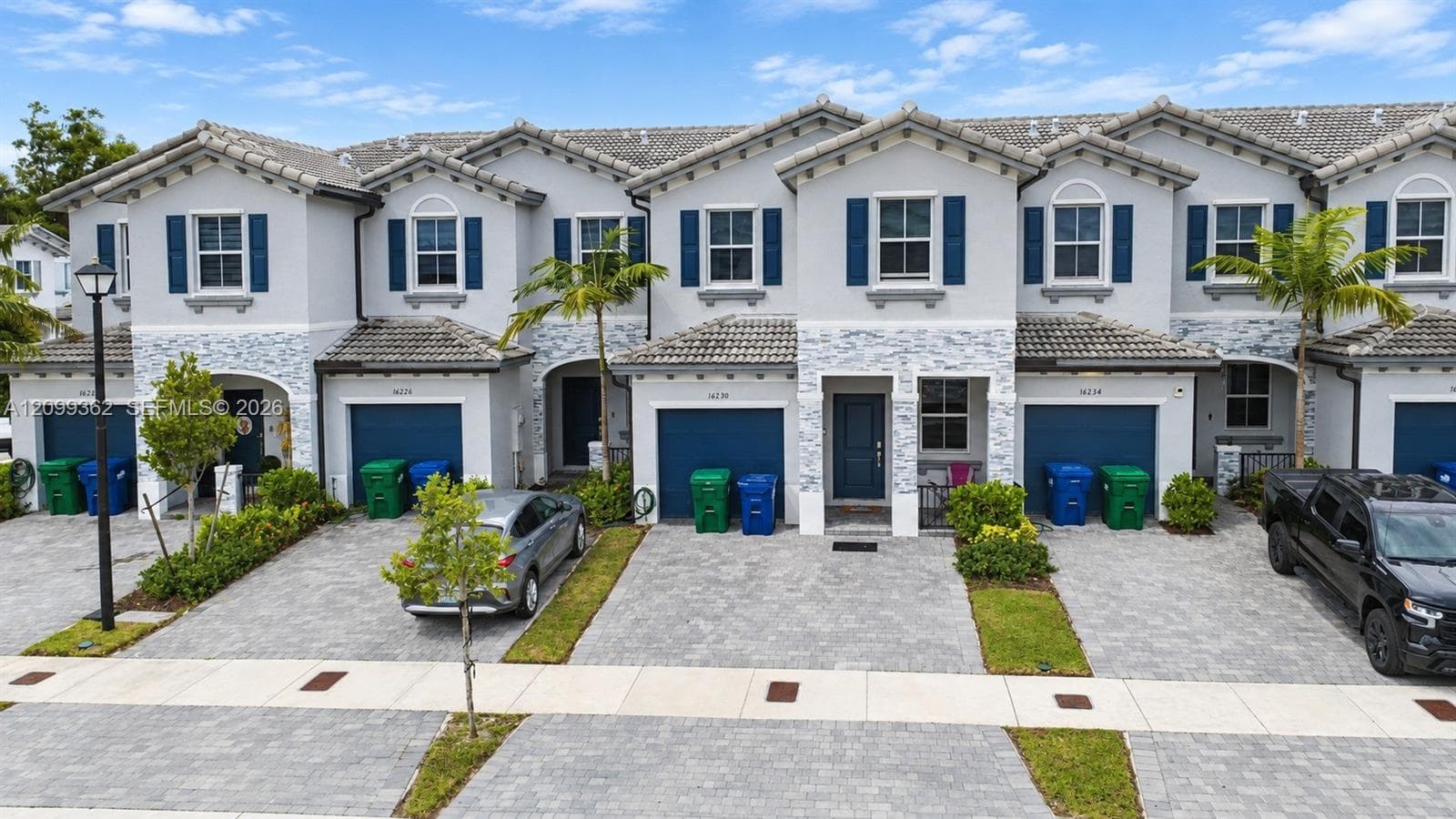 Two-story townhome exterior featuring stucco siding, stone veneer accents, multi-pane windows with decorative shutters, tile roofing, and paver driveways