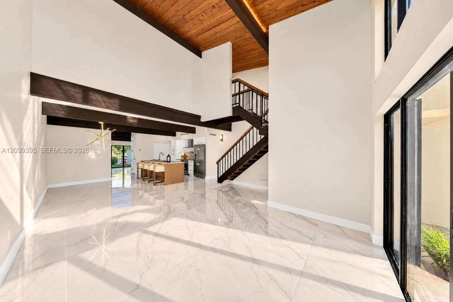 High-ceiling interior featuring a cedar wood-plank ceiling with exposed structural beams, large-format tile flooring, a geometric multi-arm light fixture, and a floating staircase with bronze metal railings