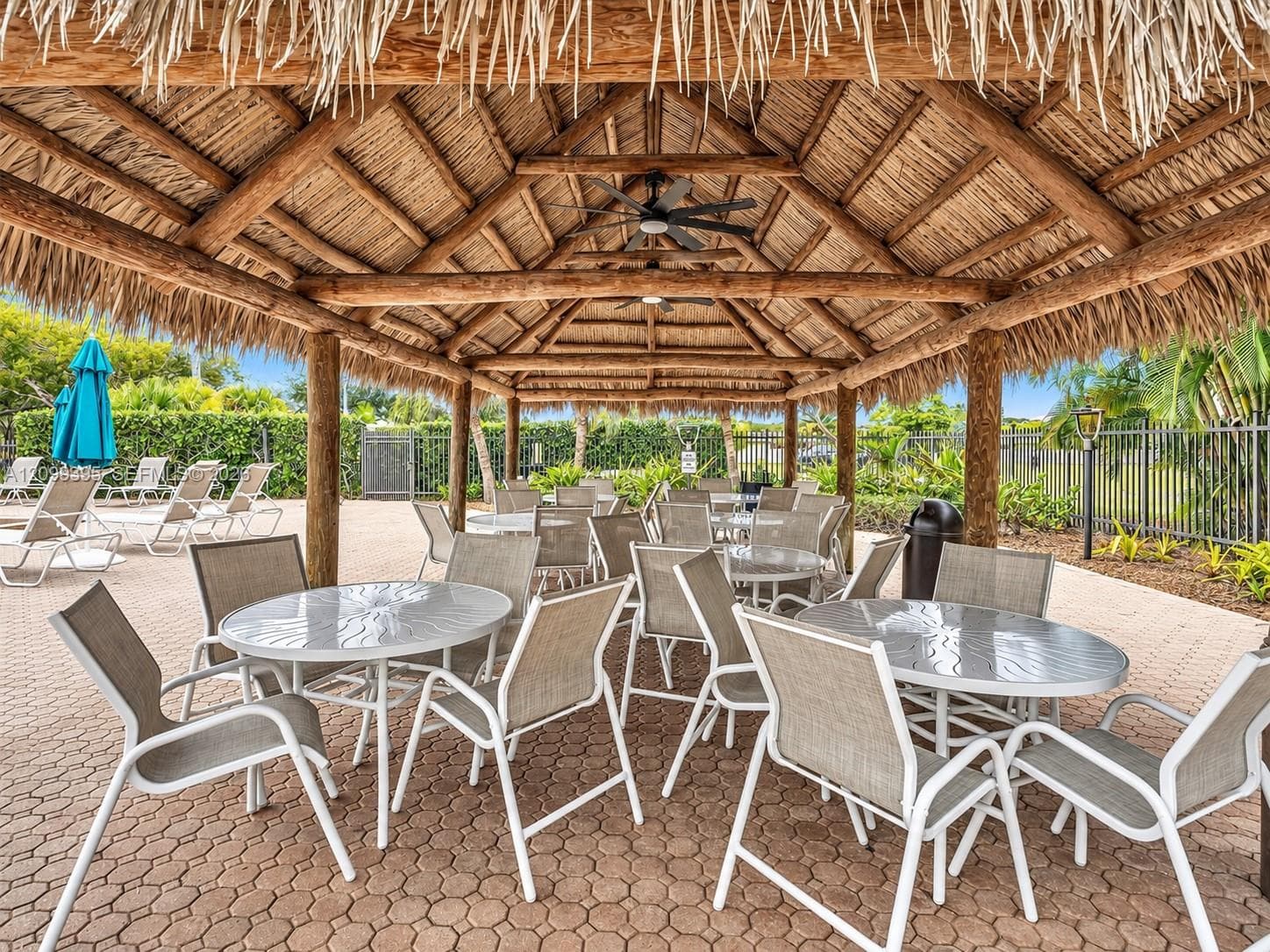 Outdoor pavilion featuring a thatched roof, exposed timber support beams, and ceiling-mounted fans over paved hardscaping