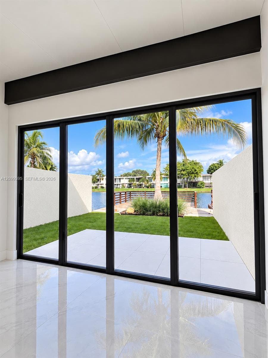 Floor-to-ceiling multi-panel sliding glass doors with matte black framing, exposed structural ceiling beam, polished stone-finish flooring, paved exterior patio, and water frontage