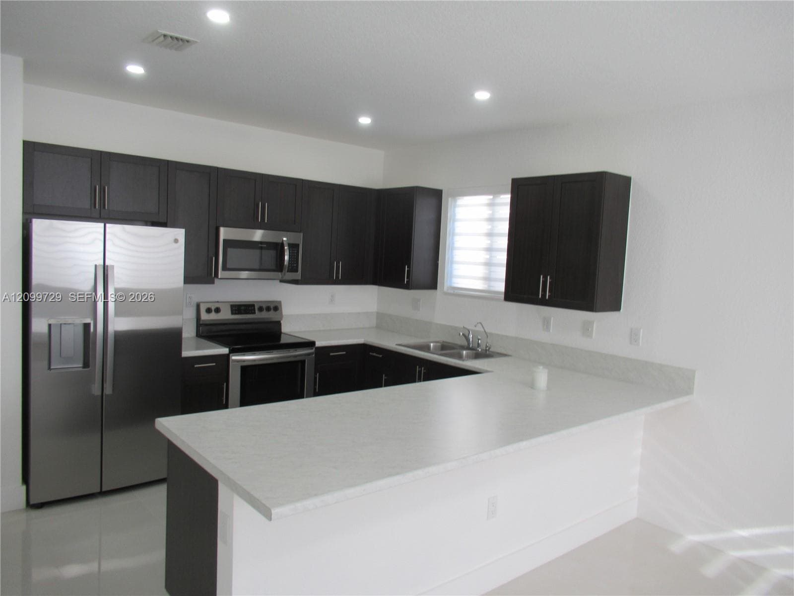 Kitchen featuring dark-toned cabinetry, stainless steel appliances, light-toned solid surface countertops, a breakfast bar, and recessed lighting