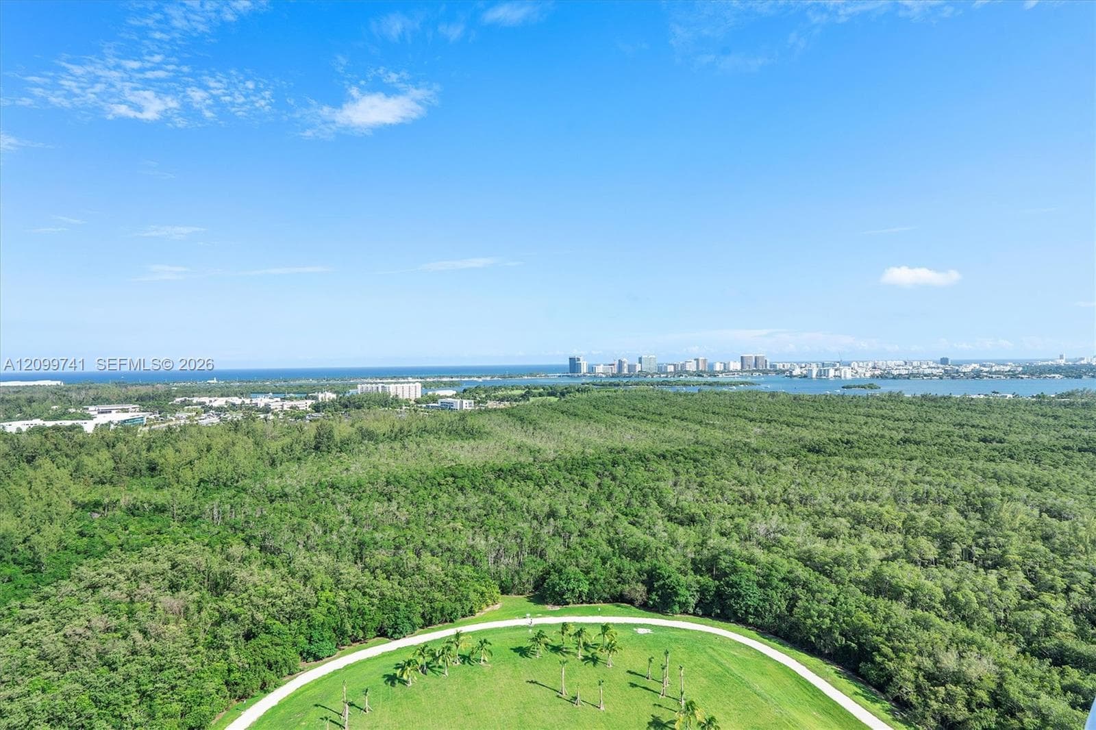 Aerial perspective of a sprawling nature preserve featuring mature tree canopy, manicured lawns, circular paved pathways, and coastal horizon