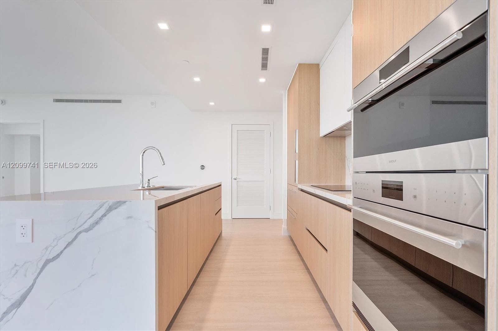 Modern kitchen featuring light-wood cabinetry, stone waterfall island, recessed ceiling lighting, and stainless steel built-in double wall ovens