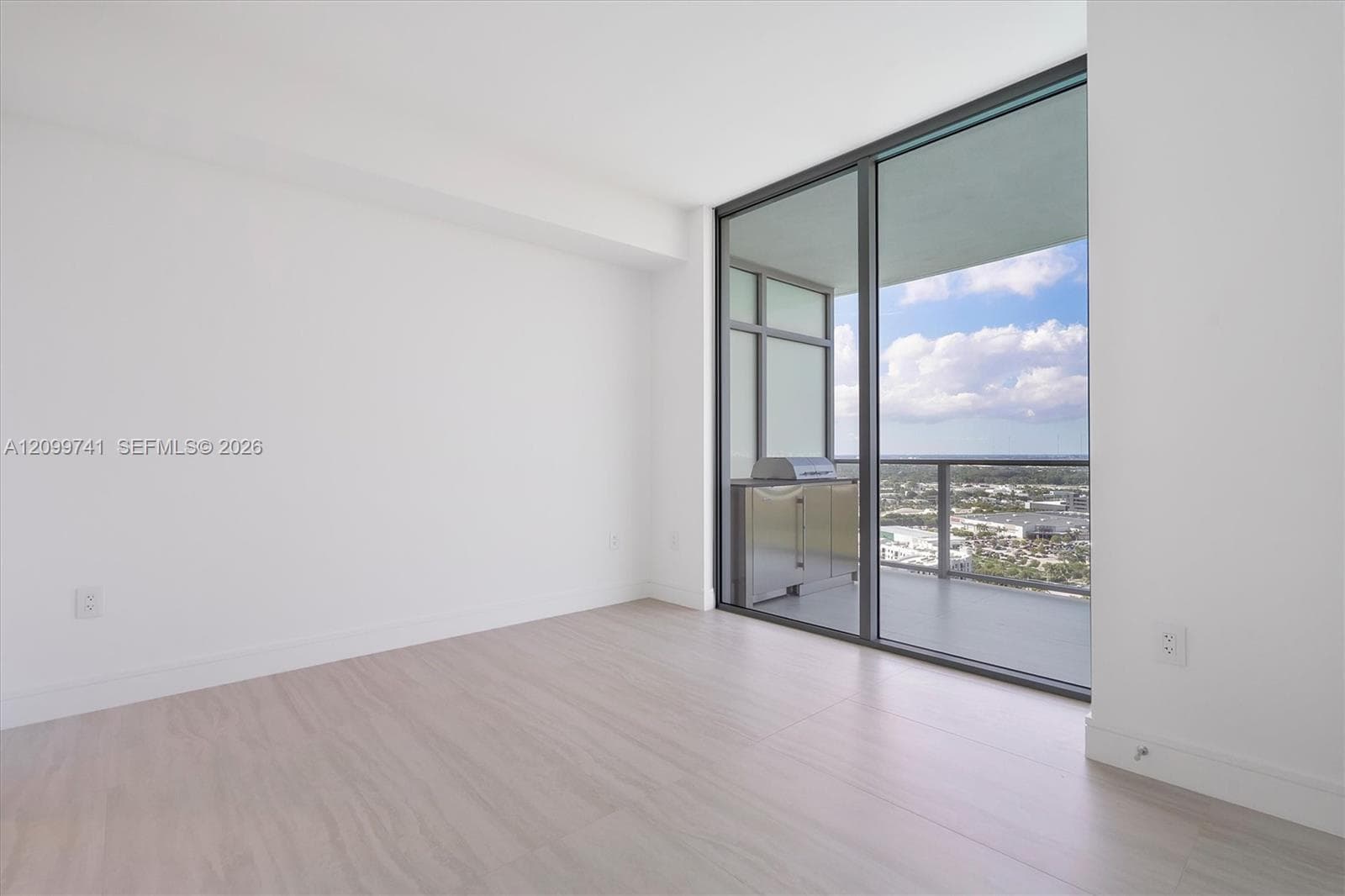 Floor-to-ceiling glass paneling, wood-finish flooring, and a covered balcony featuring a built-in outdoor kitchen with metal cabinetry