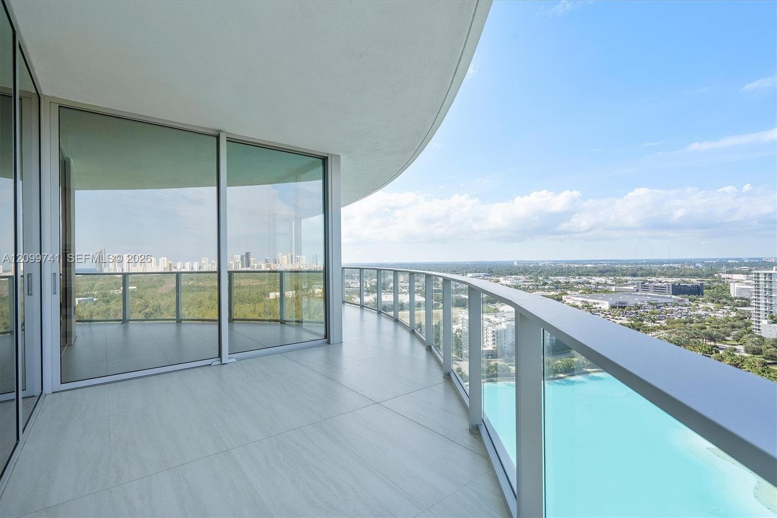Curved wrap-around balcony featuring tile flooring, glass panel railings, floor-to-ceiling sliding glass doors, and panoramic exterior exposure
