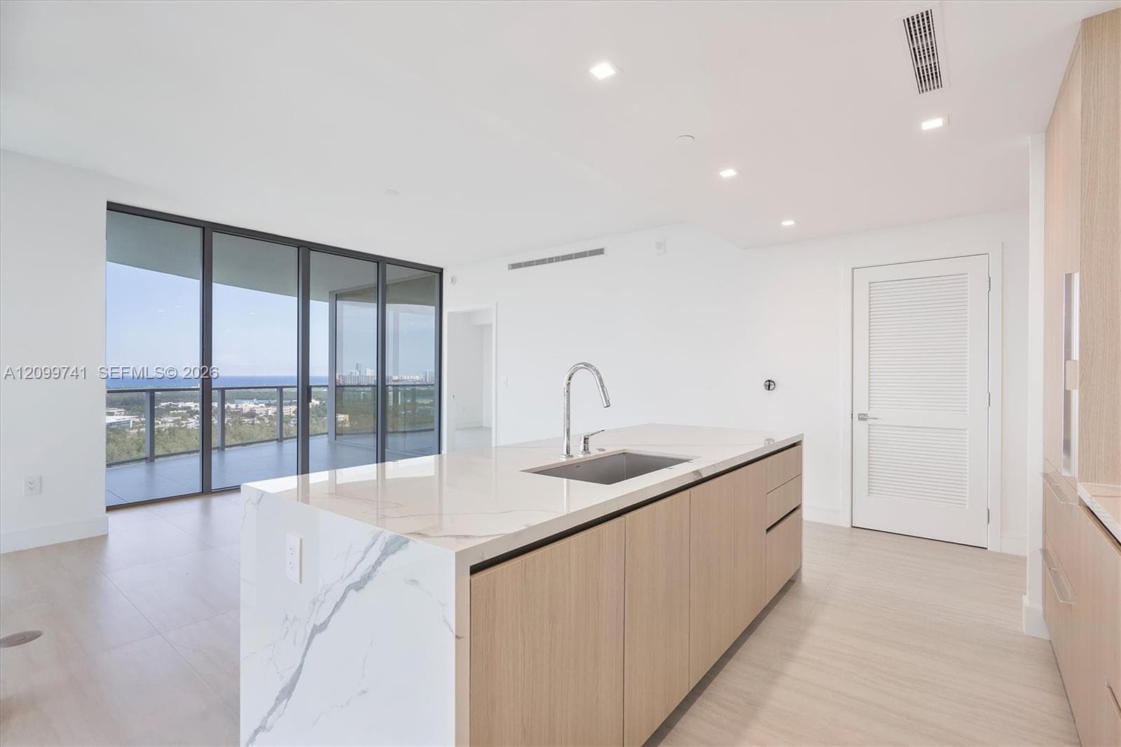 Kitchen island featuring a stone-look waterfall countertop, undermount sink with a gooseneck faucet, light wood-finish cabinetry, floor-to-ceiling glass panel walls, and recessed lighting