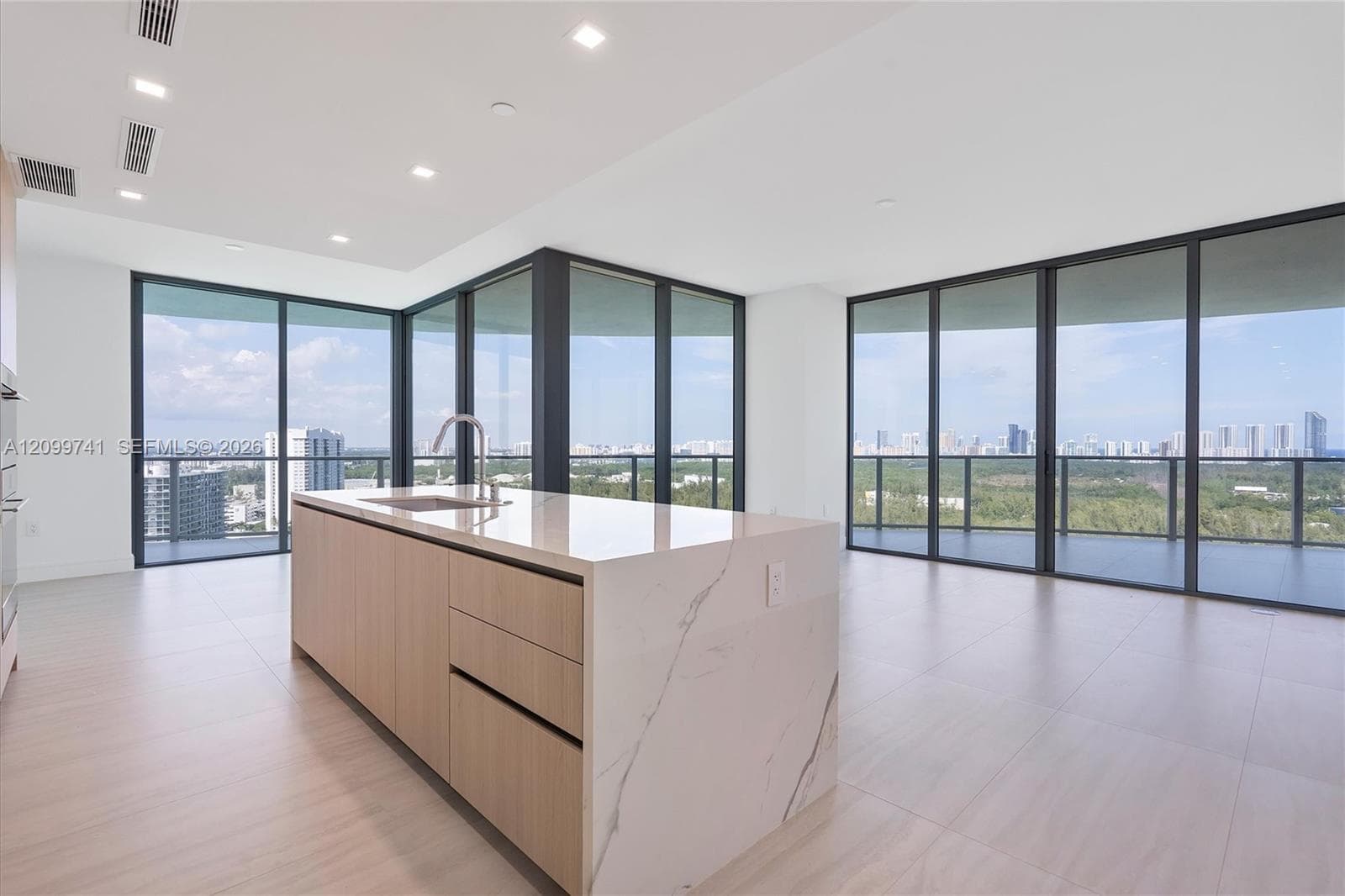 Kitchen island featuring light wood-finish cabinetry, a waterfall stone-finish countertop, and a built-in sink, illuminated by recessed lighting with floor-to-ceiling glass panel walls