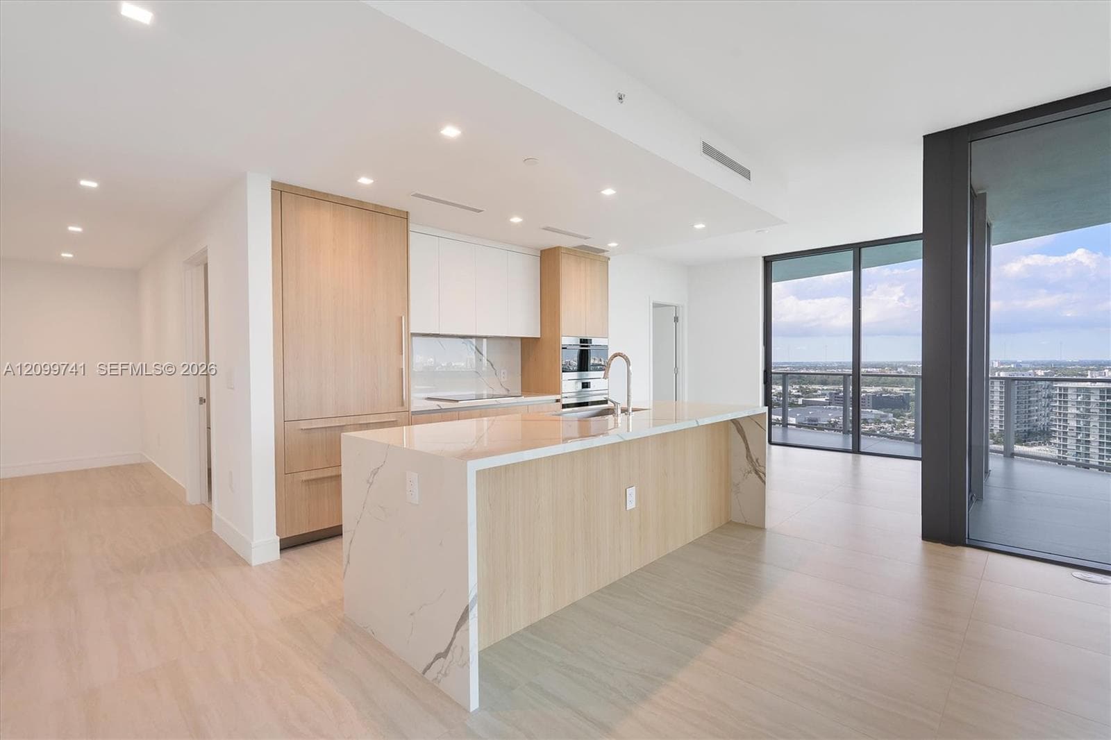 Kitchen featuring light-finish wood cabinetry, integrated appliances, stone-surface waterfall island, matte metal faucet, and floor-to-ceiling glass sliding doors