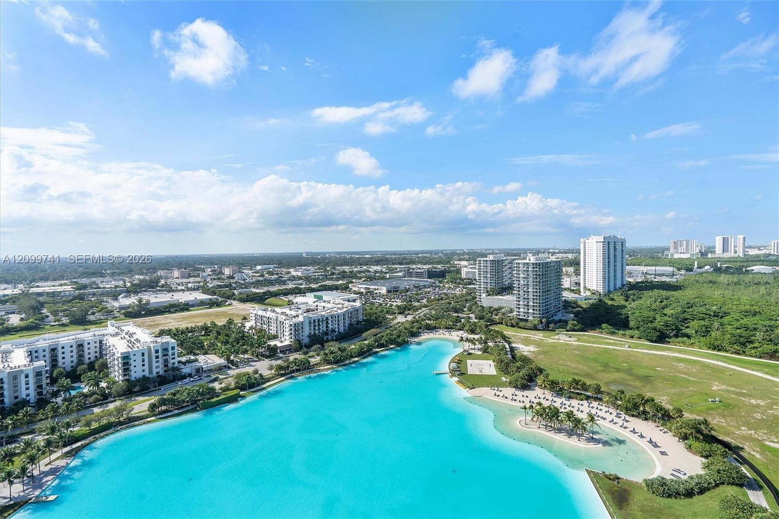 Aerial perspective of a large turquoise lagoon featuring a sandy shoreline, palm-lined beach area, and surrounding green space