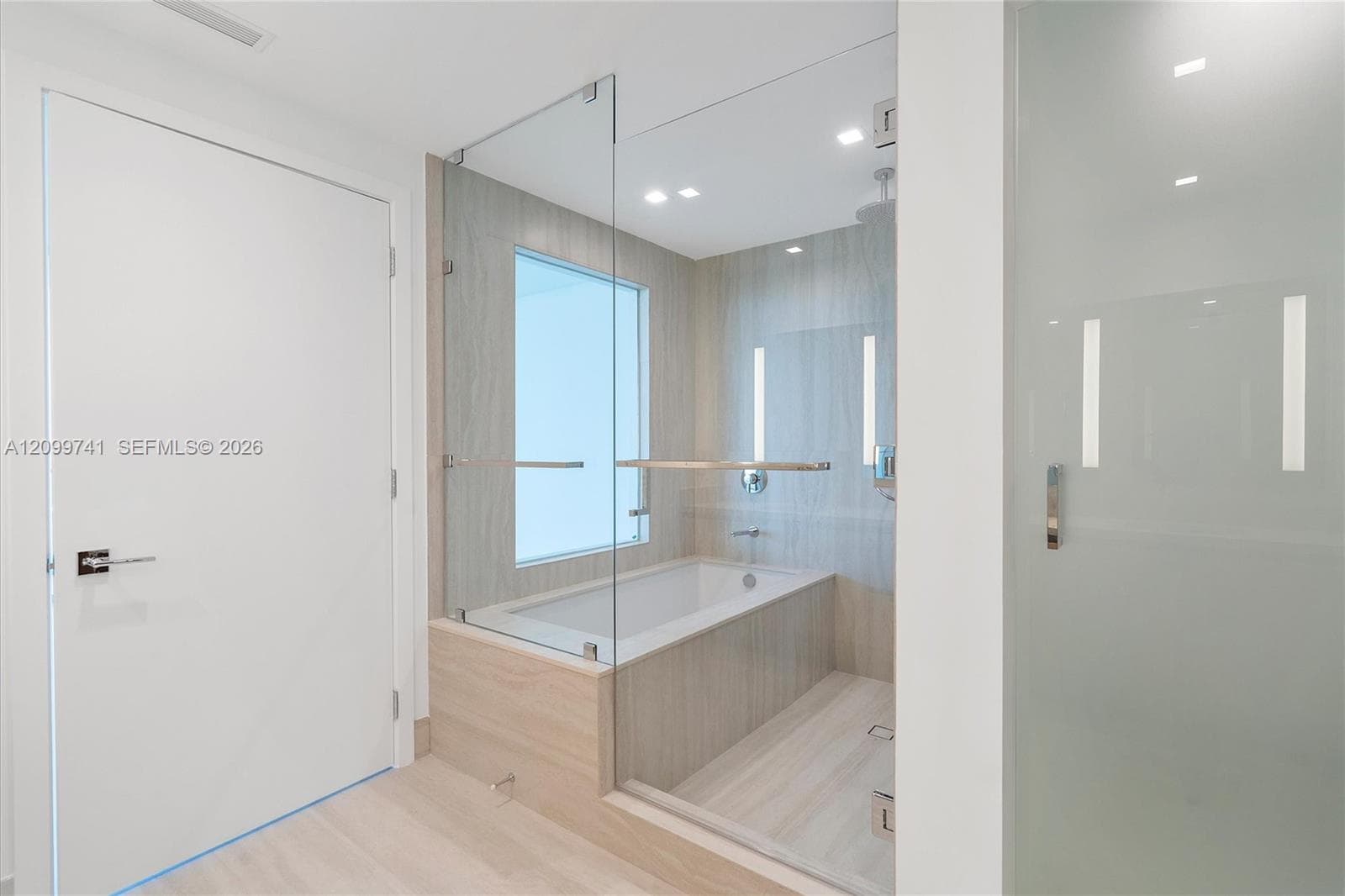 Glass-enclosed soaking tub, wall-to-wall neutral tile surround, recessed ceiling lighting, and frosted privacy door