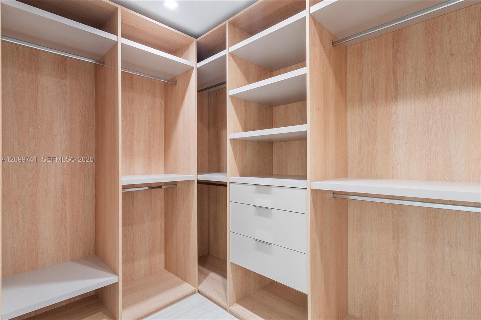 Custom walk-in closet featuring light wood-finish cabinetry, built-in open shelving, tiered hanging rods, and a three-drawer storage unit with minimalist hardware