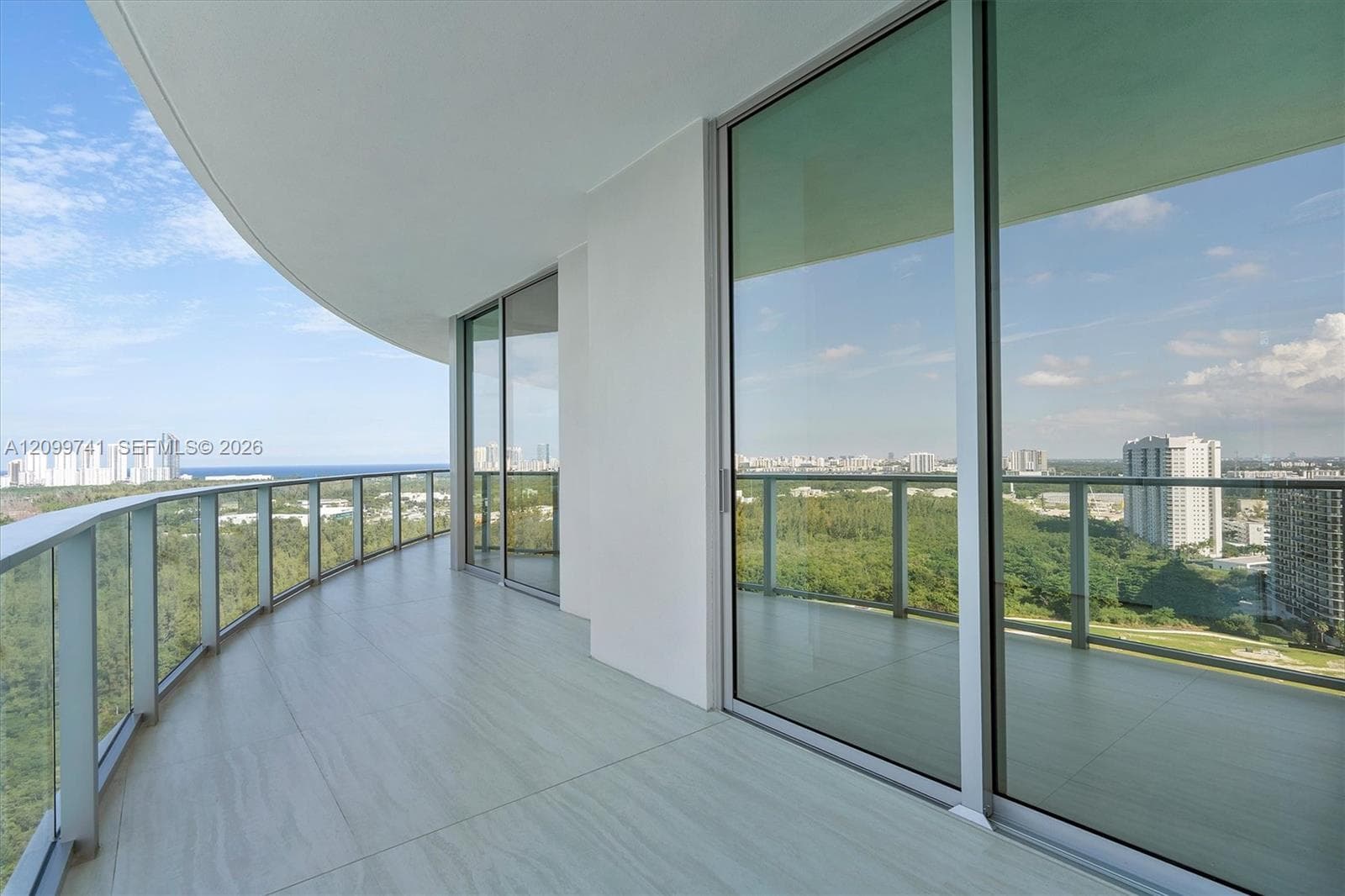Curved balcony featuring large-format stone-finish tile flooring, a transparent glass panel railing, and floor-to-ceiling sliding glass doors