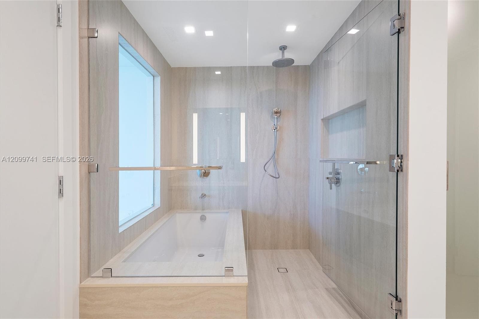 Walk-in glass shower with integrated wall niche and ceiling-mounted rain showerhead, complemented by a deep soaking tub featuring neutral stone-finish wall tile and recessed lighting