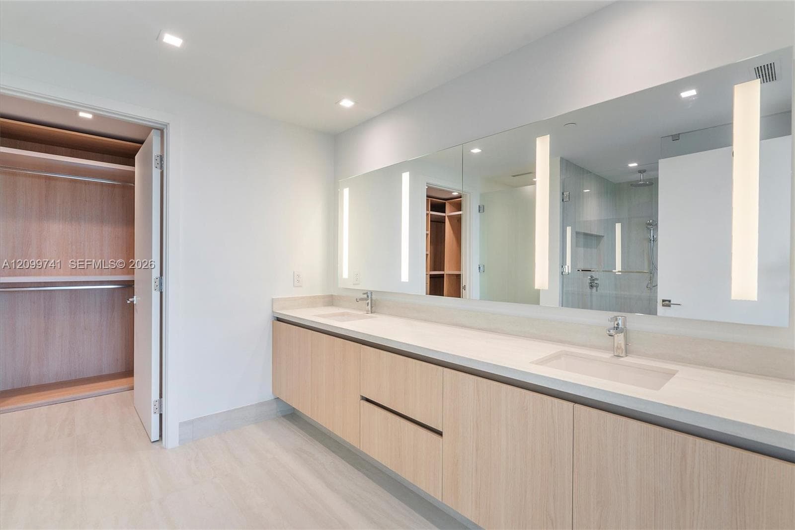 Wall-mounted light-wood vanity with dual rectangular undermount sinks and solid-surface countertop featuring integrated vertical lighting within a large mirror, recessed ceiling lighting, and a walk-in closet with built-in wooden shelving