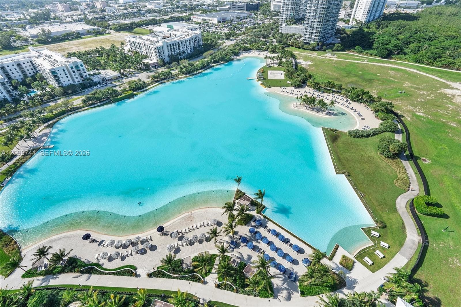 Aerial perspective of a large lagoon featuring sandy shorelines, paved pathways, manicured green space, and mature palm trees
