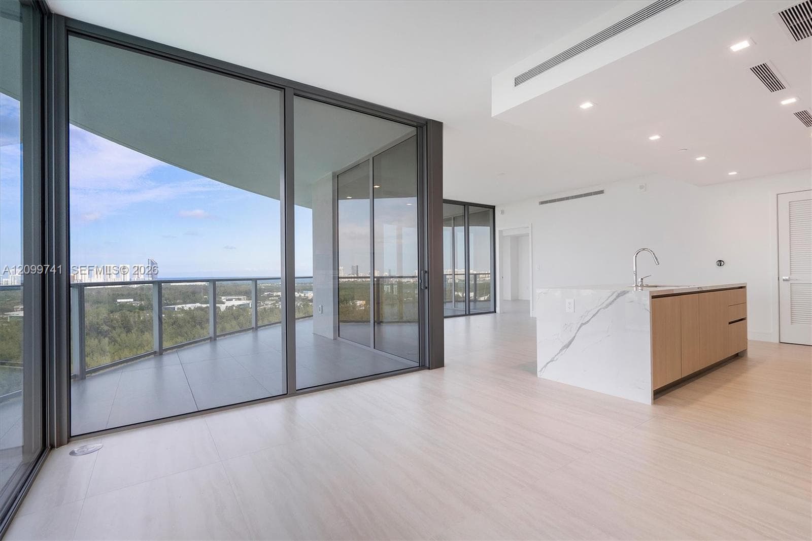 Floor-to-ceiling glass sliding doors opening to a wraparound balcony, kitchen island with waterfall-edge countertop, wood-finish cabinetry, and recessed lighting