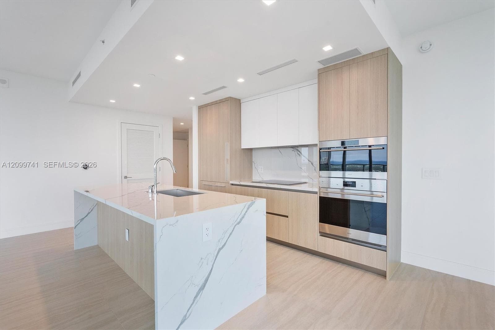 Contemporary kitchen featuring an island with a waterfall-edge countertop, stone-patterned backsplash, light wood-finish cabinetry, built-in stainless steel double ovens, and recessed lighting
