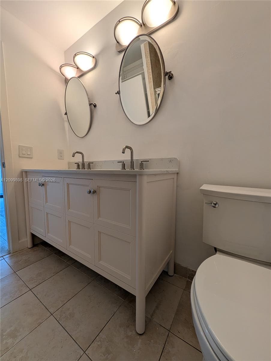 Dual-sink vanity featuring a stone countertop, oval wall-mounted mirrors, brushed-nickel fixtures, and tile flooring