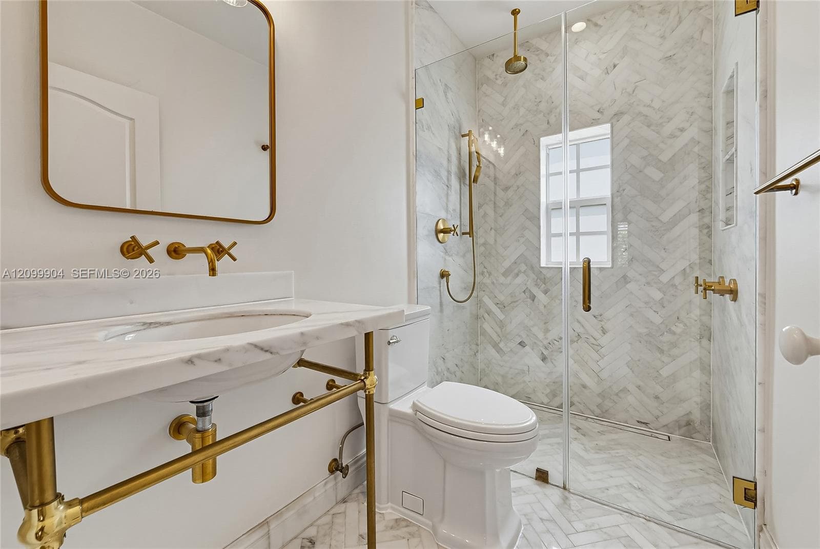 Wall-mounted brass faucets, marble-surround vanity with integrated sink, walk-in shower with herringbone tile, brass hardware, and glass shower enclosure