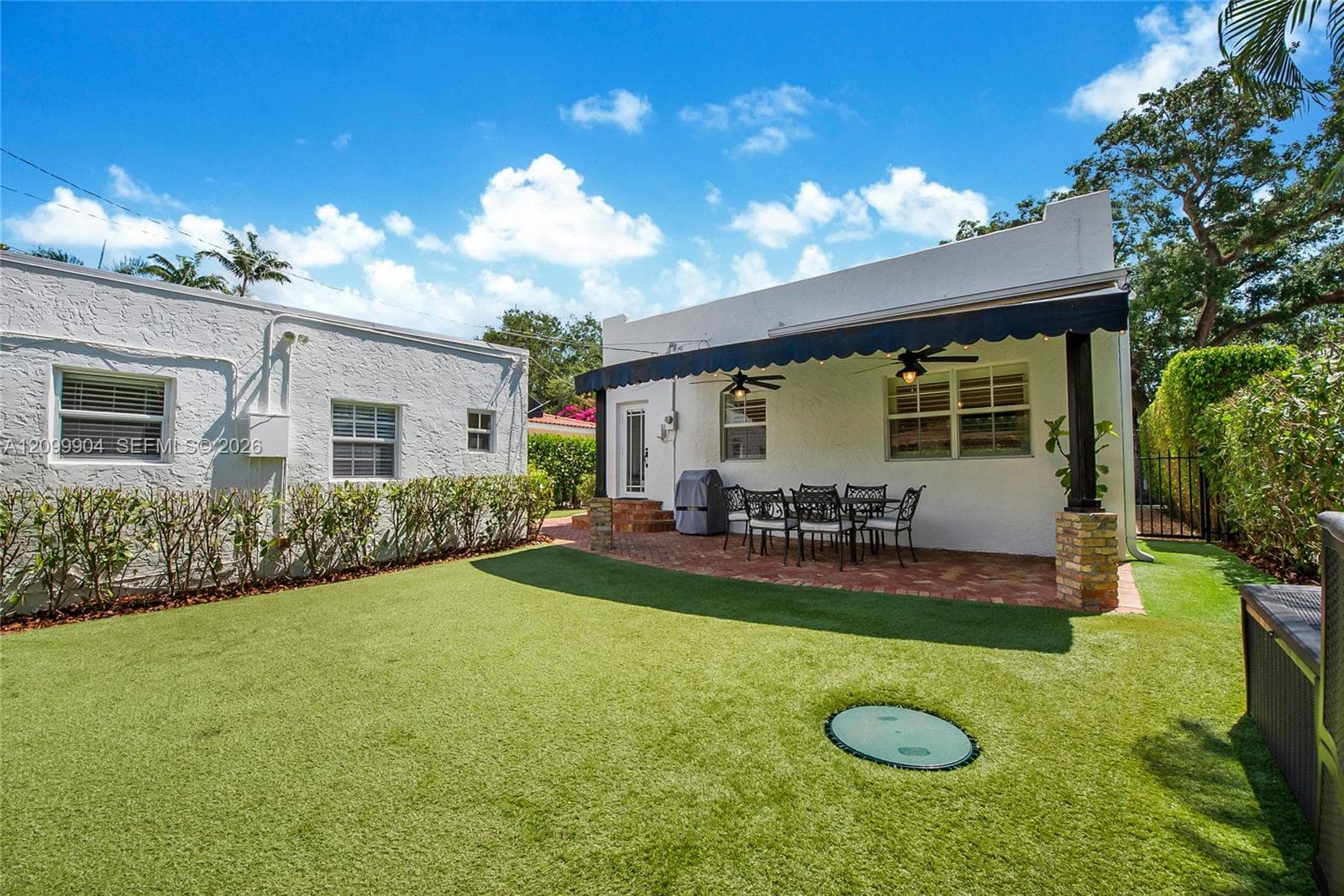 Rear exterior featuring a brick-paved patio, retractable scalloped awning, and stone-accented support columns, finished with a synthetic turf lawn and manicured perimeter hedges