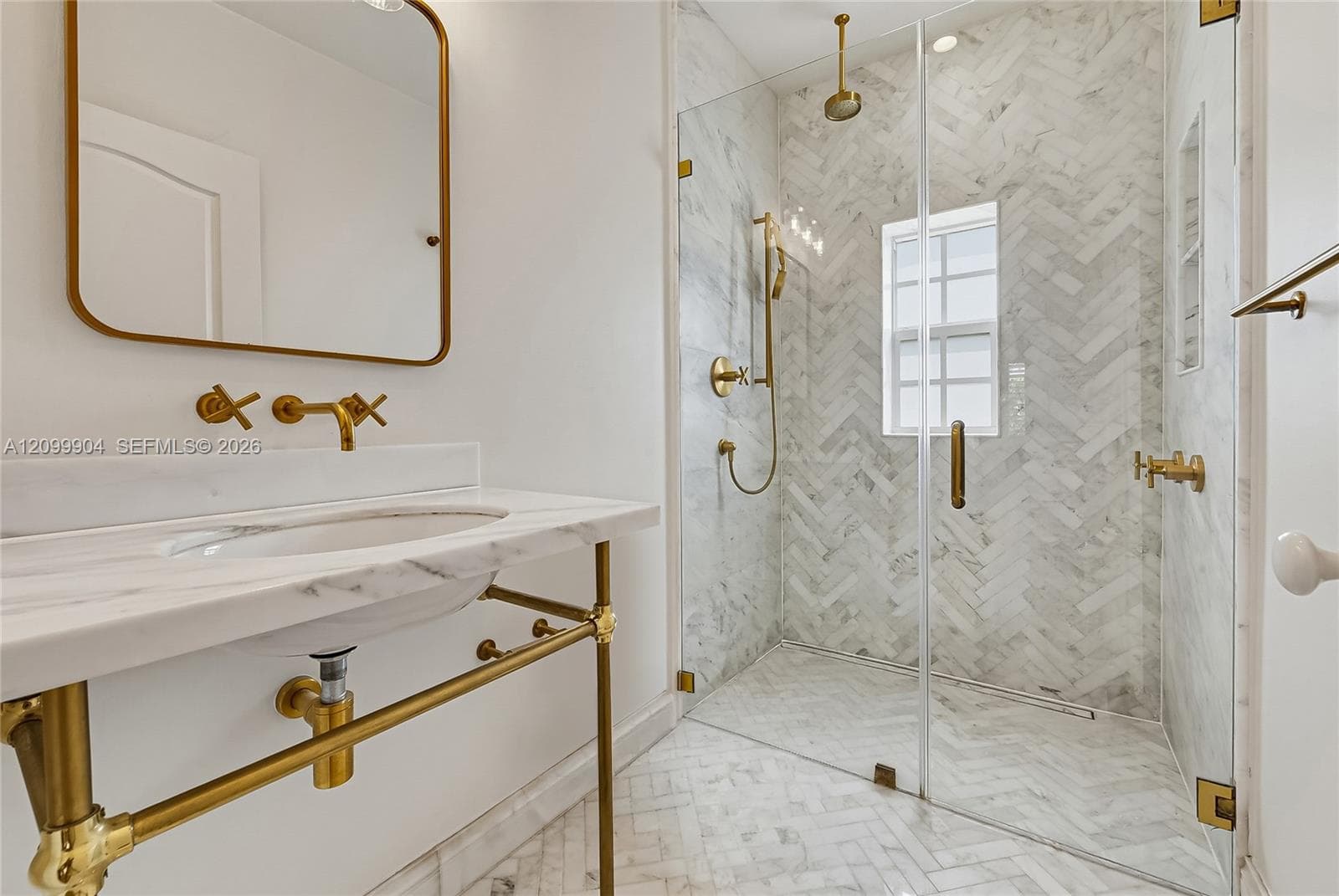 Console-style vanity with a stone countertop and brass-finish plumbing fixtures, glass-enclosed walk-in shower featuring herringbone tile pattern, and a brass-framed wall mirror
